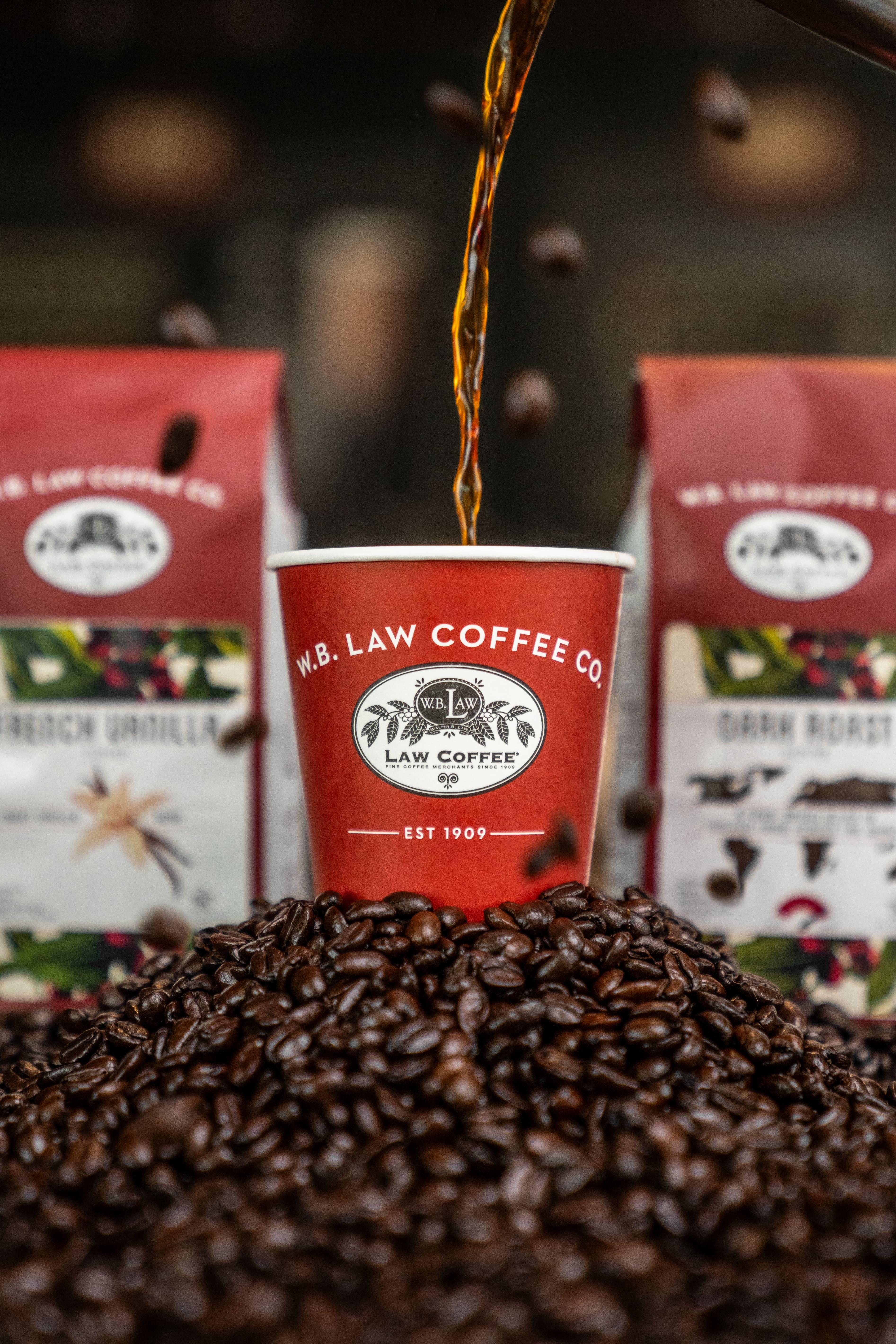 Red cup with 'W.B. Law Coffee Co.' logo being filled with coffee, surrounded by coffee beans and bags.