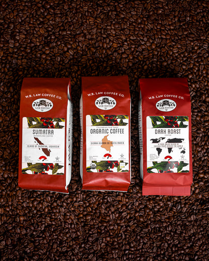 Three bags of dark roasted WB Law Coffees displayed on a background of coffee beans. Each bag has a red label with white text indicating the type of coffee, such as 'Sumatra, Dark Roast, and Organic Colombian Coffee'.