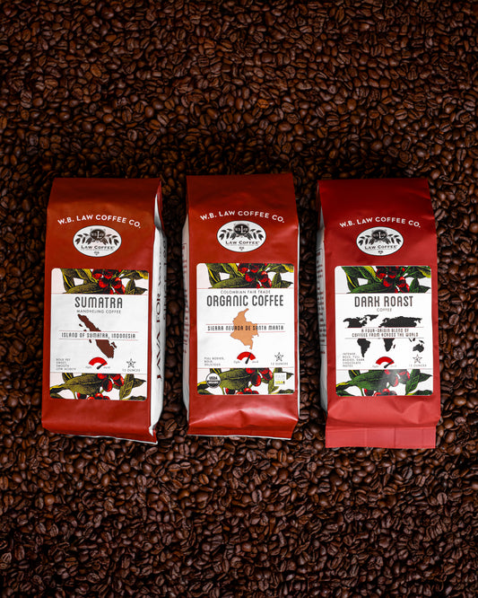 Three bags of dark roasted WB Law Coffees displayed on a background of coffee beans. Each bag has a red label with white text indicating the type of coffee, such as 'Sumatra, Dark Roast, and Organic Colombian Coffee'.