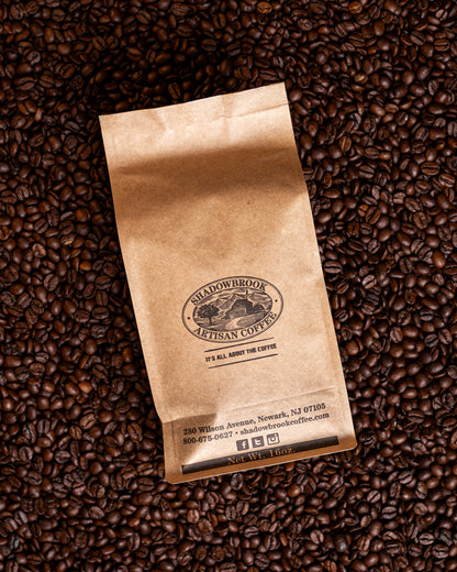 A brown bag of Shadowbrook Artisan Coffee beans sitting on pile of roasted coffee beans. 