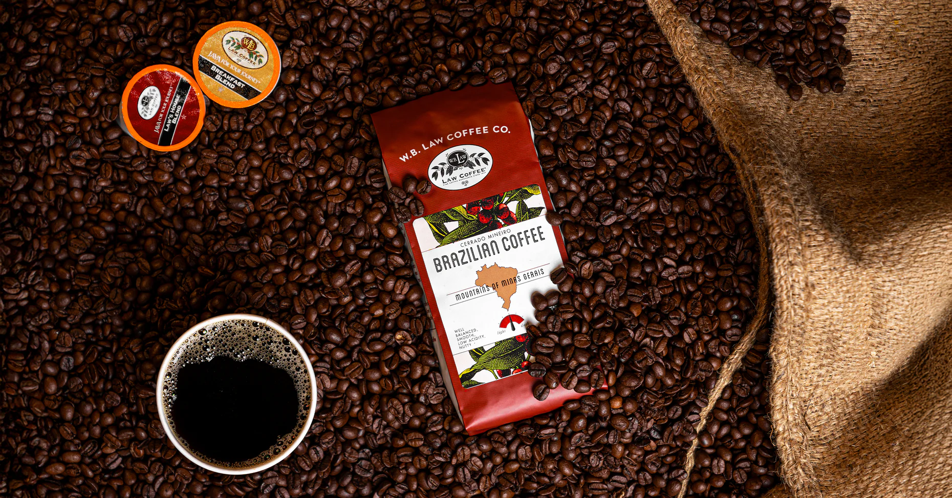 WB Law Coffee Co. Brazilian Cerrado coffee bag sitting next to a cup of black coffee and two kcups in a pile of coffee beans.