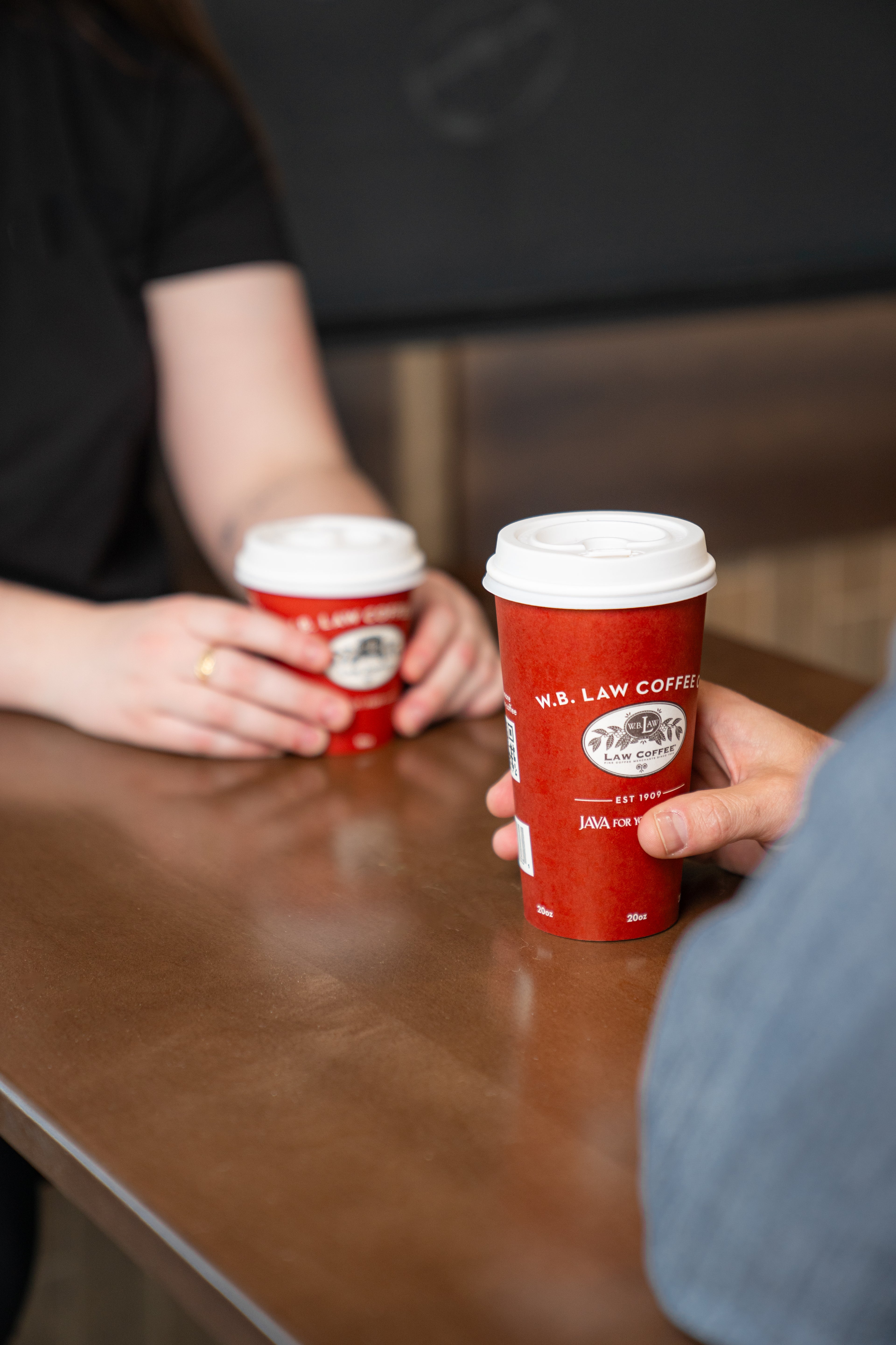 Two WB Law Coffee coffee cups with white lids on a wooden table, one held by a person wearing a black shirt and the other by a person wearing a gray sweater.