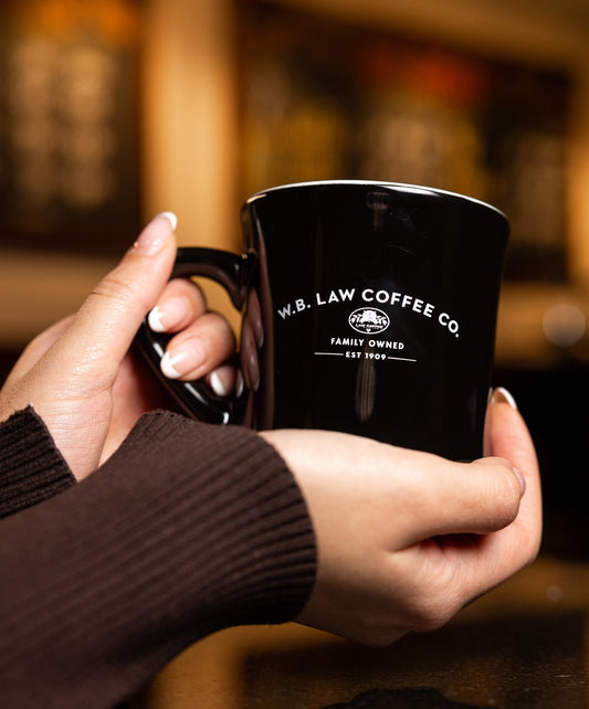 Black mug with 'W.B. Law Coffee Co.' branding held by a person in a blurred coffee shop setting.